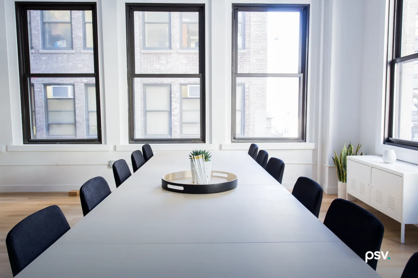 An empty conference room with a long white table, a row of dark chairs down each side, a black tray holding a cup of pens, three tall black framed windows looking onto a brick building across the street, a plant and a low white cabinet. An empty conference room with a long white table, a row of dark chairs down each side, a black tray holding a cup of pens, three tall black framed windows looking onto a brick building across the street, a plant and a low white cabinet.