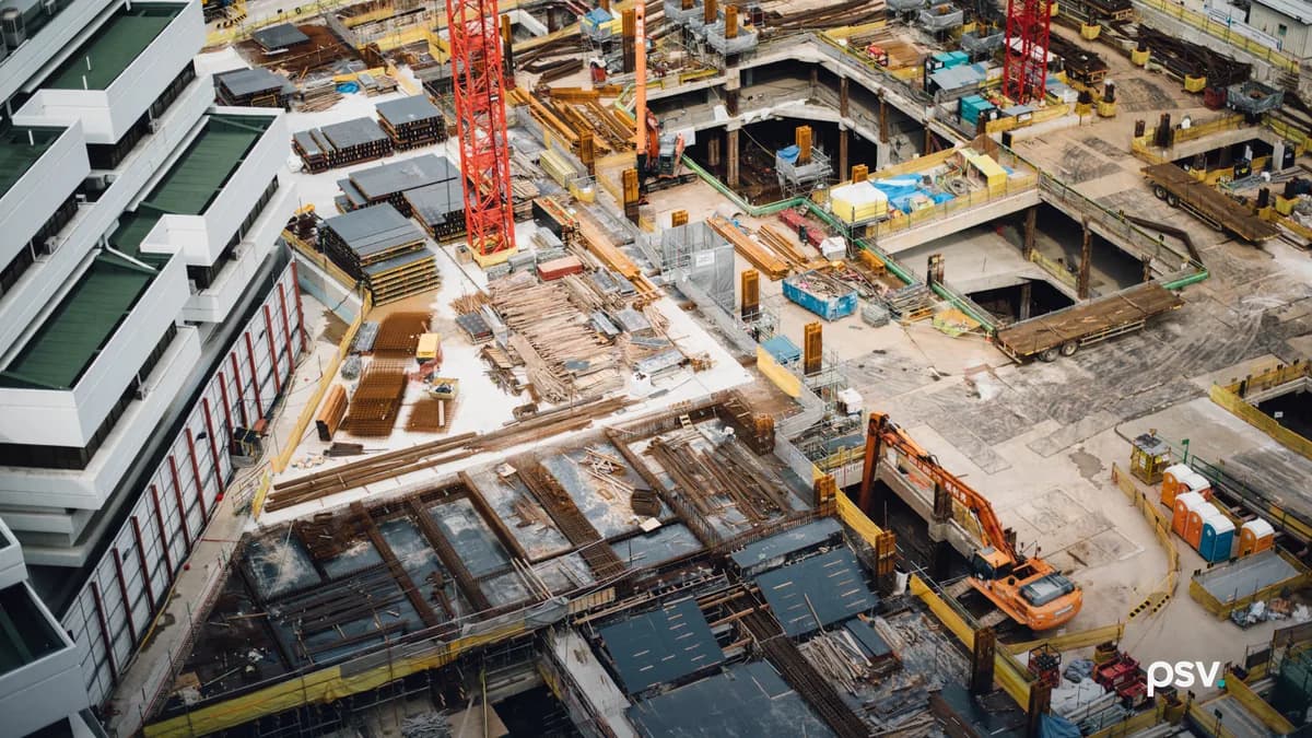 An aerial view of a large urban construction site showing rebar mats, stacked formwork panels, timber shoring, two red tower crane masts, orange excavators, portable toilets and open basement decks beside a finished white building.