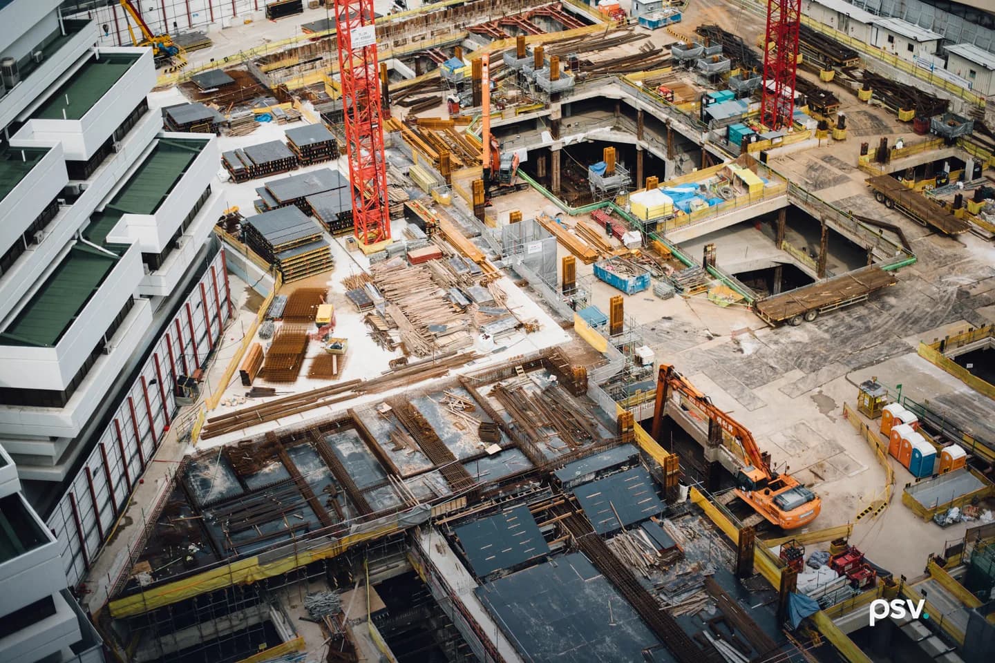 An aerial view of a large urban construction site showing rebar mats, stacked formwork panels, timber shoring, two red tower crane masts, orange excavators, portable toilets and open basement decks beside a finished white building. An aerial view of a large urban construction site showing rebar mats, stacked formwork panels, timber shoring, two red tower crane masts, orange excavators, portable toilets and open basement decks beside a finished white building.
