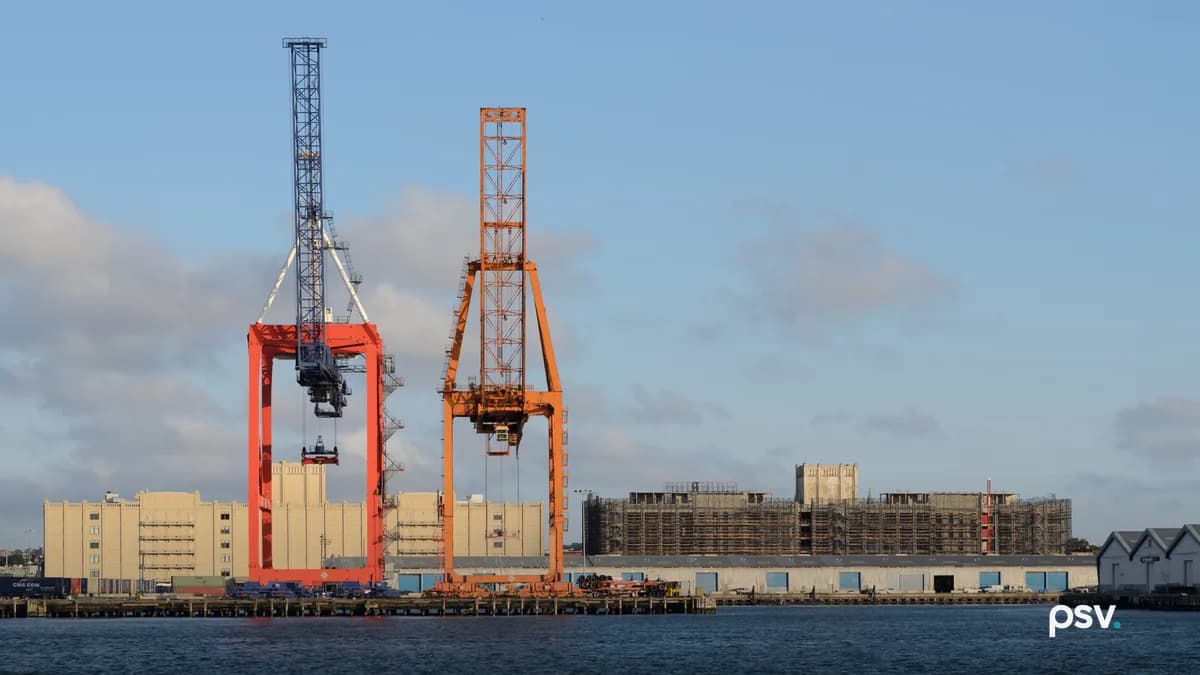 Two gantry cranes, one orange and one blue, stand over a container terminal wharf on the far side of open water in low sun, with stacked containers, low warehouse sheds and a scaffolded building along the pier behind them.