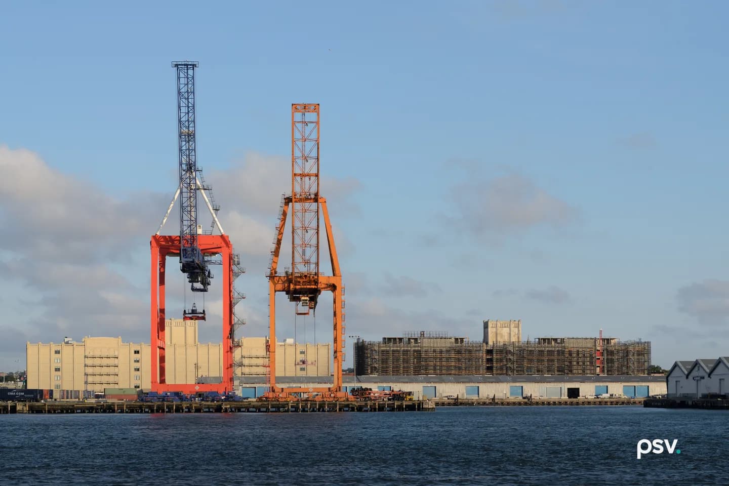 Two gantry cranes, one orange and one blue, stand over a container terminal wharf on the far side of open water in low sun, with stacked containers, low warehouse sheds and a scaffolded building along the pier behind them. Two gantry cranes, one orange and one blue, stand over a container terminal wharf on the far side of open water in low sun, with stacked containers, low warehouse sheds and a scaffolded building along the pier behind them.