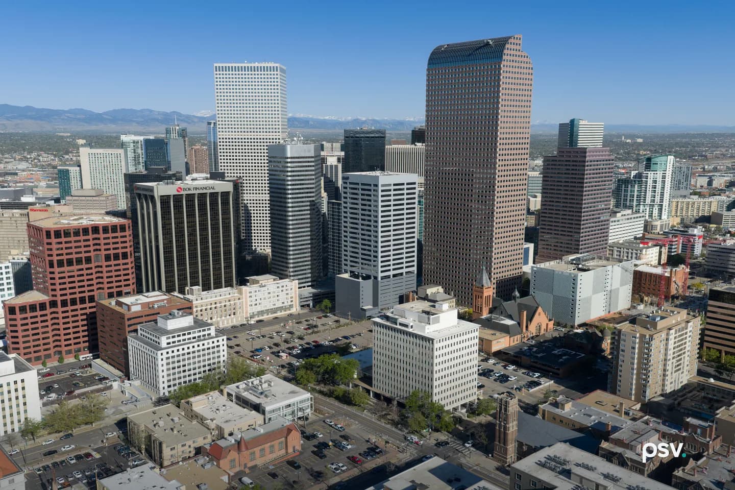 An aerial view over downtown Denver on a clear day, showing clusters of glass and stone office towers, lower brick buildings, surface parking lots and the street grid below, with mountains on the distant horizon.