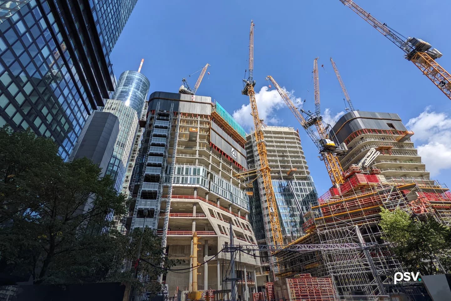 Looking up from street level at several high-rise towers under construction in Frankfurt's financial district: exposed concrete slabs and columns, climbing formwork, safety screens, partly installed glass curtain wall, and tower cranes against a blue sky. Looking up from street level at several high-rise towers under construction in Frankfurt's financial district: exposed concrete slabs and columns, climbing formwork, safety screens, partly installed glass curtain wall, and tower cranes against a blue sky.