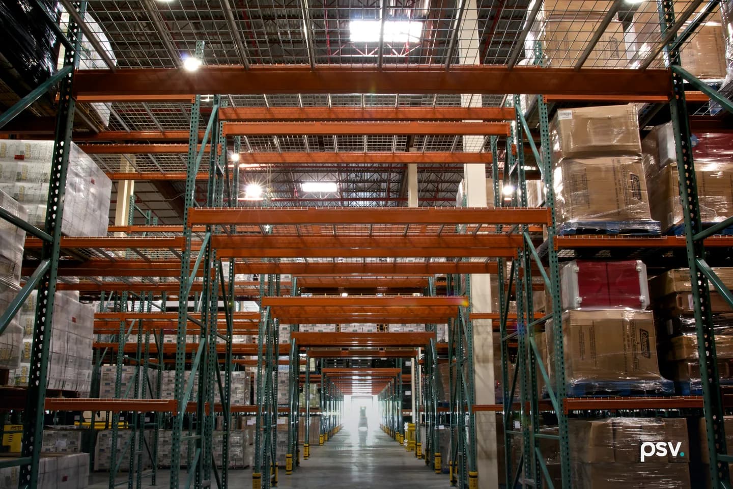 Interior of a distribution warehouse: tall steel pallet racking in orange and green runs down both sides of a wide concrete aisle stacked with shrink-wrapped pallets and cartons, receding toward a lit doorway at the far end. Interior of a distribution warehouse: tall steel pallet racking in orange and green runs down both sides of a wide concrete aisle stacked with shrink-wrapped pallets and cartons, receding toward a lit doorway at the far end.
