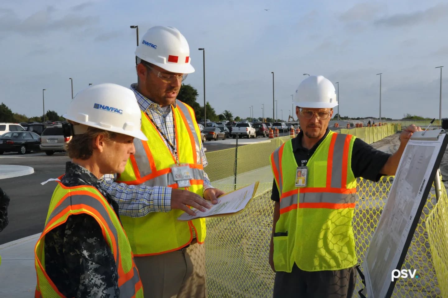 Three people in yellow high-visibility vests and white hard hats stand at the edge of an active construction site. The man at center reads from a paper report while the man at right steadies a large drawing sheet on a board, with green site fencing, orange cones and graded ground behind them. Three people in yellow high-visibility vests and white hard hats stand at the edge of an active construction site. The man at center reads from a paper report while the man at right steadies a large drawing sheet on a board, with green site fencing, orange cones and graded ground behind them.