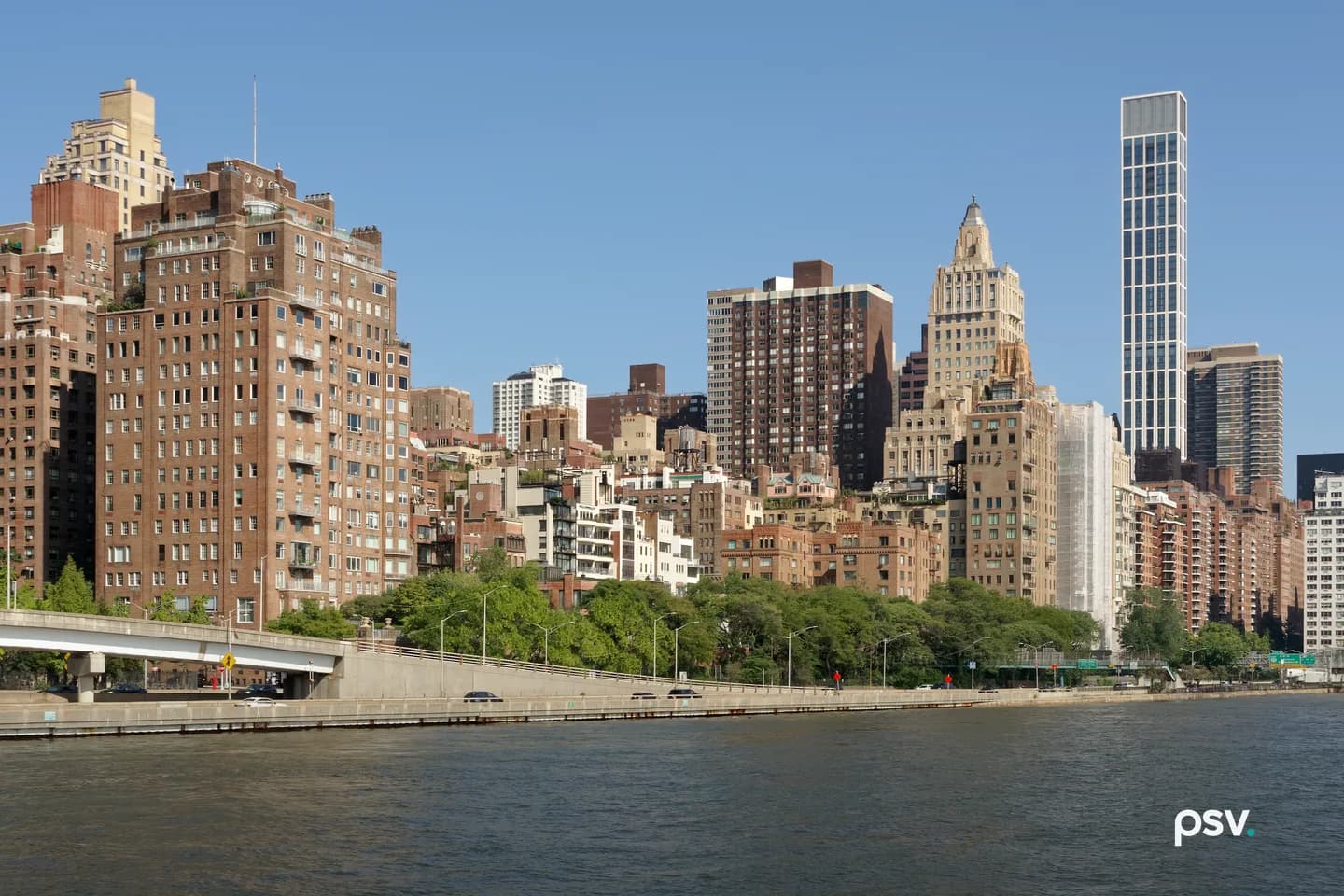 Row of brick and stone apartment towers along the Manhattan bank of the East River on a clear day, with a slim modern high-rise at right, trees and a highway ramp below, and river water in the foreground. Row of brick and stone apartment towers along the Manhattan bank of the East River on a clear day, with a slim modern high-rise at right, trees and a highway ramp below, and river water in the foreground.