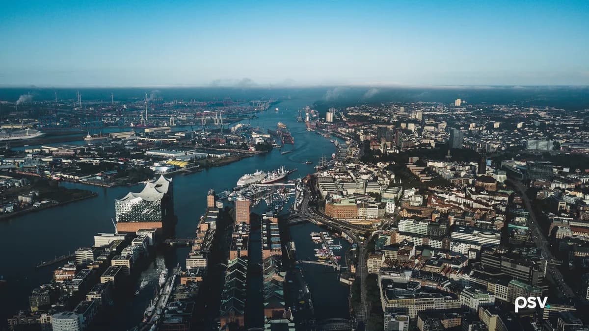 Aerial daylight view over Hamburg's harbour district: brick warehouse blocks with copper-green roofs, a glass concert hall, modern office buildings, a canal of moored boats, and container cranes lining the far bank of the Elbe.