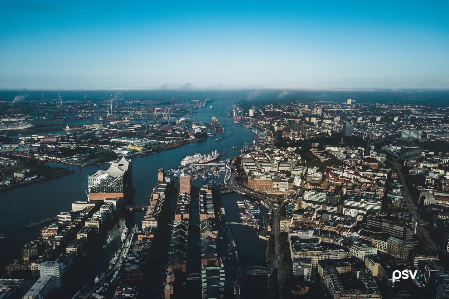 Aerial daylight view over Hamburg's harbour district: brick warehouse blocks with copper-green roofs, a glass concert hall, modern office buildings, a canal of moored boats, and container cranes lining the far bank of the Elbe. Aerial daylight view over Hamburg's harbour district: brick warehouse blocks with copper-green roofs, a glass concert hall, modern office buildings, a canal of moored boats, and container cranes lining the far bank of the Elbe.