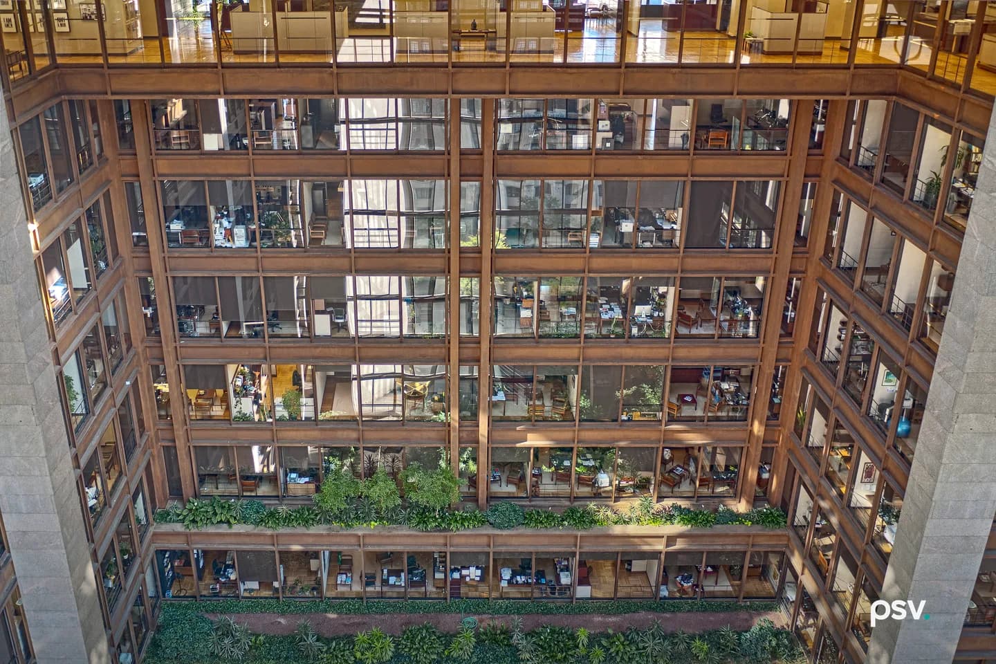 Looking across the planted indoor atrium of the Ford Foundation building in New York, where tiers of glass-walled offices holding desks, screens and paperwork stack above terraces of dense greenery behind a rust-brown steel and teak frame. Looking across the planted indoor atrium of the Ford Foundation building in New York, where tiers of glass-walled offices holding desks, screens and paperwork stack above terraces of dense greenery behind a rust-brown steel and teak frame.