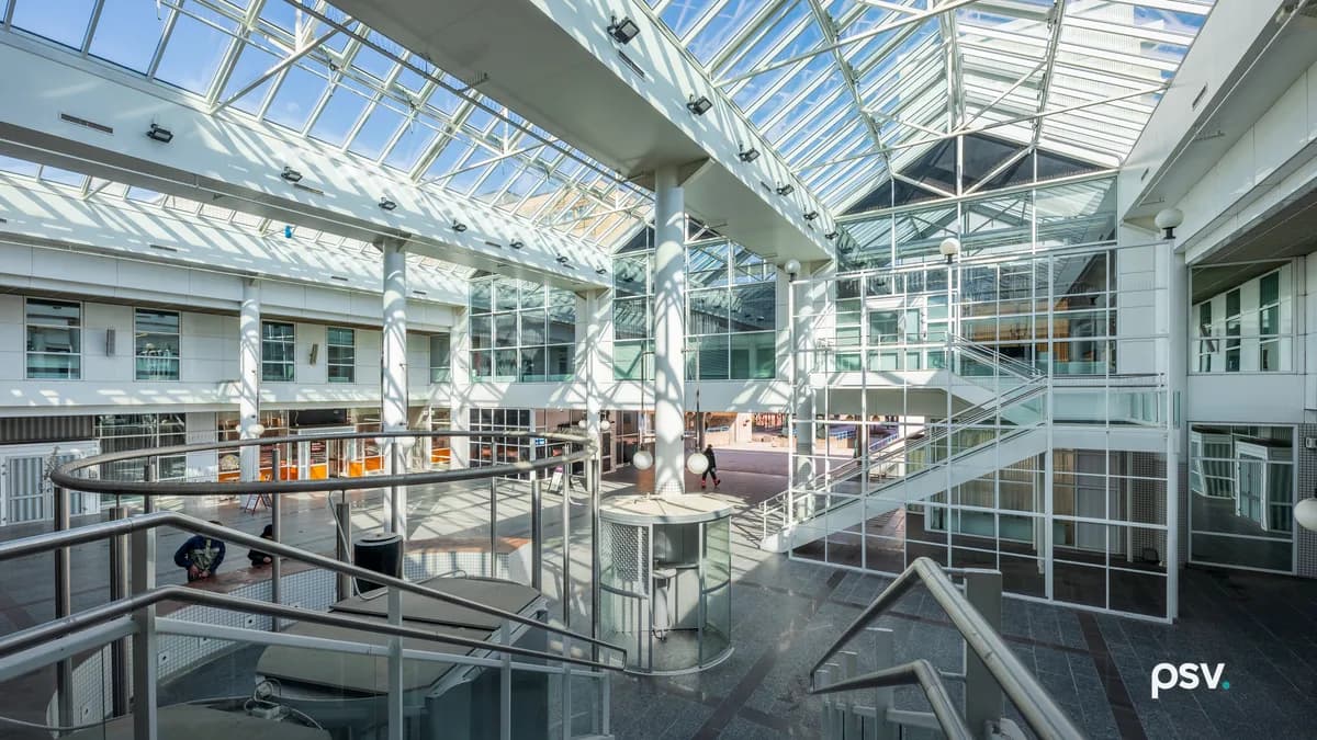A glass-roofed atrium inside an office building, with white steel trusses carrying angled skylights, balconied upper floors of windows on both sides, metal stair railings in the foreground and a polished stone floor below.