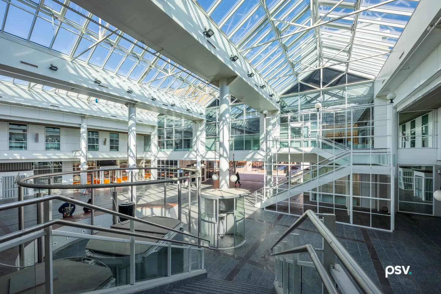 A glass-roofed atrium inside an office building, with white steel trusses carrying angled skylights, balconied upper floors of windows on both sides, metal stair railings in the foreground and a polished stone floor below. A glass-roofed atrium inside an office building, with white steel trusses carrying angled skylights, balconied upper floors of windows on both sides, metal stair railings in the foreground and a polished stone floor below.