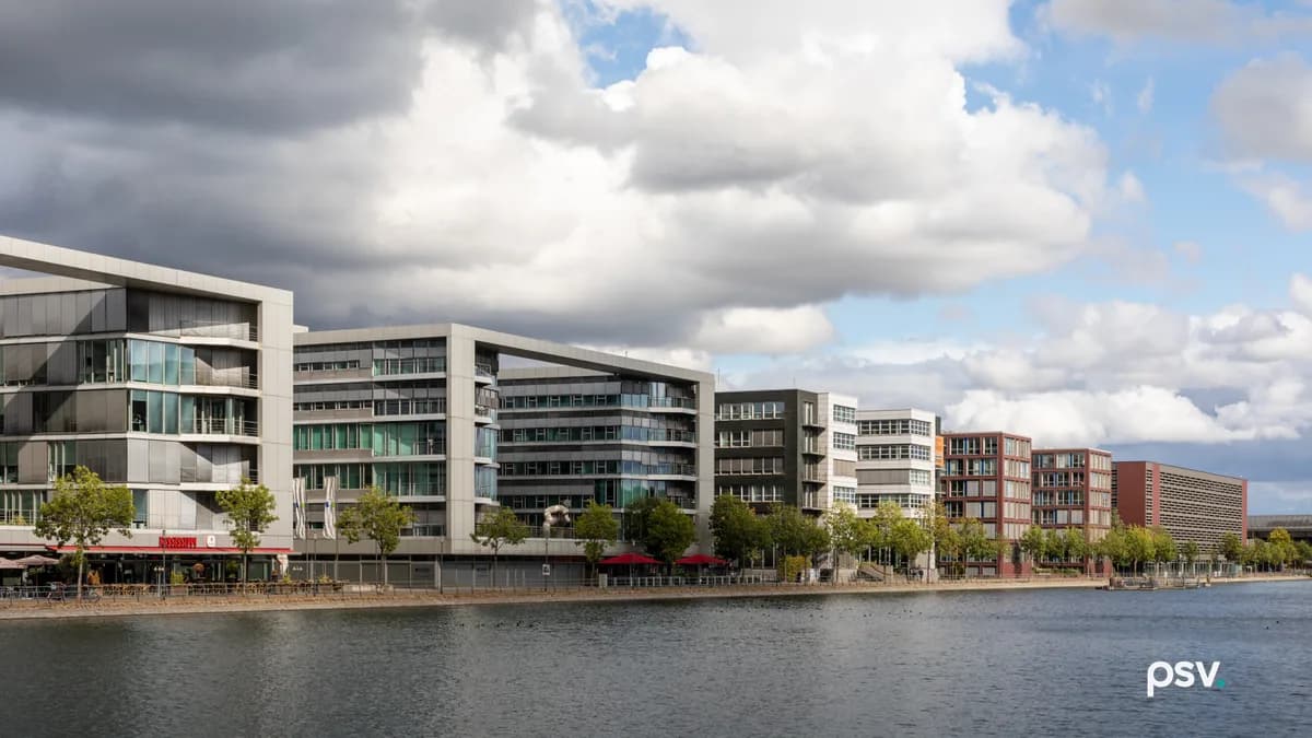 A row of modern office buildings five to seven storeys high, in silver, white, dark grey and red brick, standing along a stone-edged harbour quay under a cloudy sky, with young trees at the waterline and open water in the foreground.
