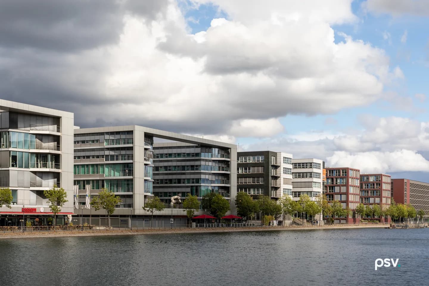 A row of modern office buildings five to seven storeys high, in silver, white, dark grey and red brick, standing along a stone-edged harbour quay under a cloudy sky, with young trees at the waterline and open water in the foreground. A row of modern office buildings five to seven storeys high, in silver, white, dark grey and red brick, standing along a stone-edged harbour quay under a cloudy sky, with young trees at the waterline and open water in the foreground.