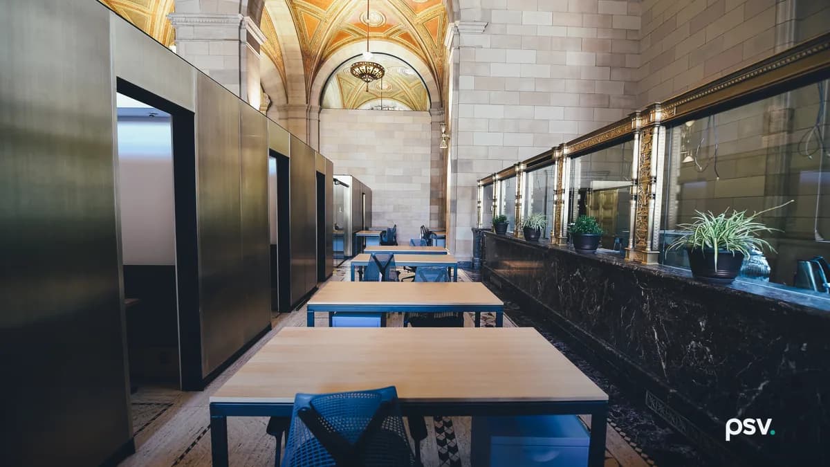 A long interior corridor in a converted heritage bank building lined with plain wood desks, blue task chairs and rolling file cabinets, with bronze-clad booths on the left, brass-framed glazed teller screens on the right, and a gilded vaulted ceiling overhead.