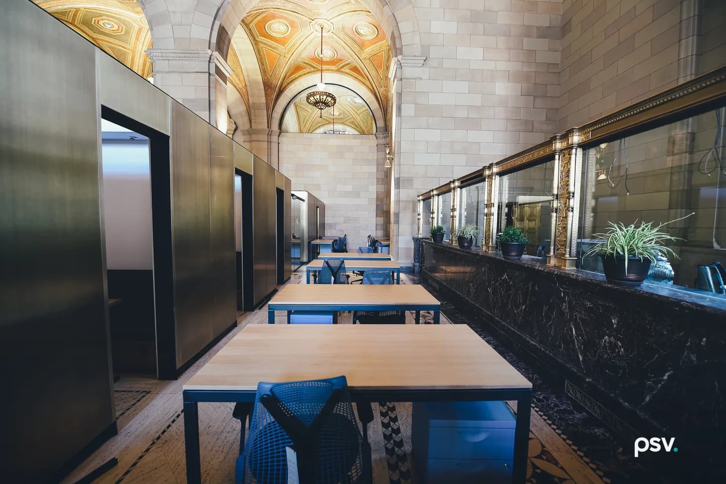 A long interior corridor in a converted heritage bank building lined with plain wood desks, blue task chairs and rolling file cabinets, with bronze-clad booths on the left, brass-framed glazed teller screens on the right, and a gilded vaulted ceiling overhead. A long interior corridor in a converted heritage bank building lined with plain wood desks, blue task chairs and rolling file cabinets, with bronze-clad booths on the left, brass-framed glazed teller screens on the right, and a gilded vaulted ceiling overhead.