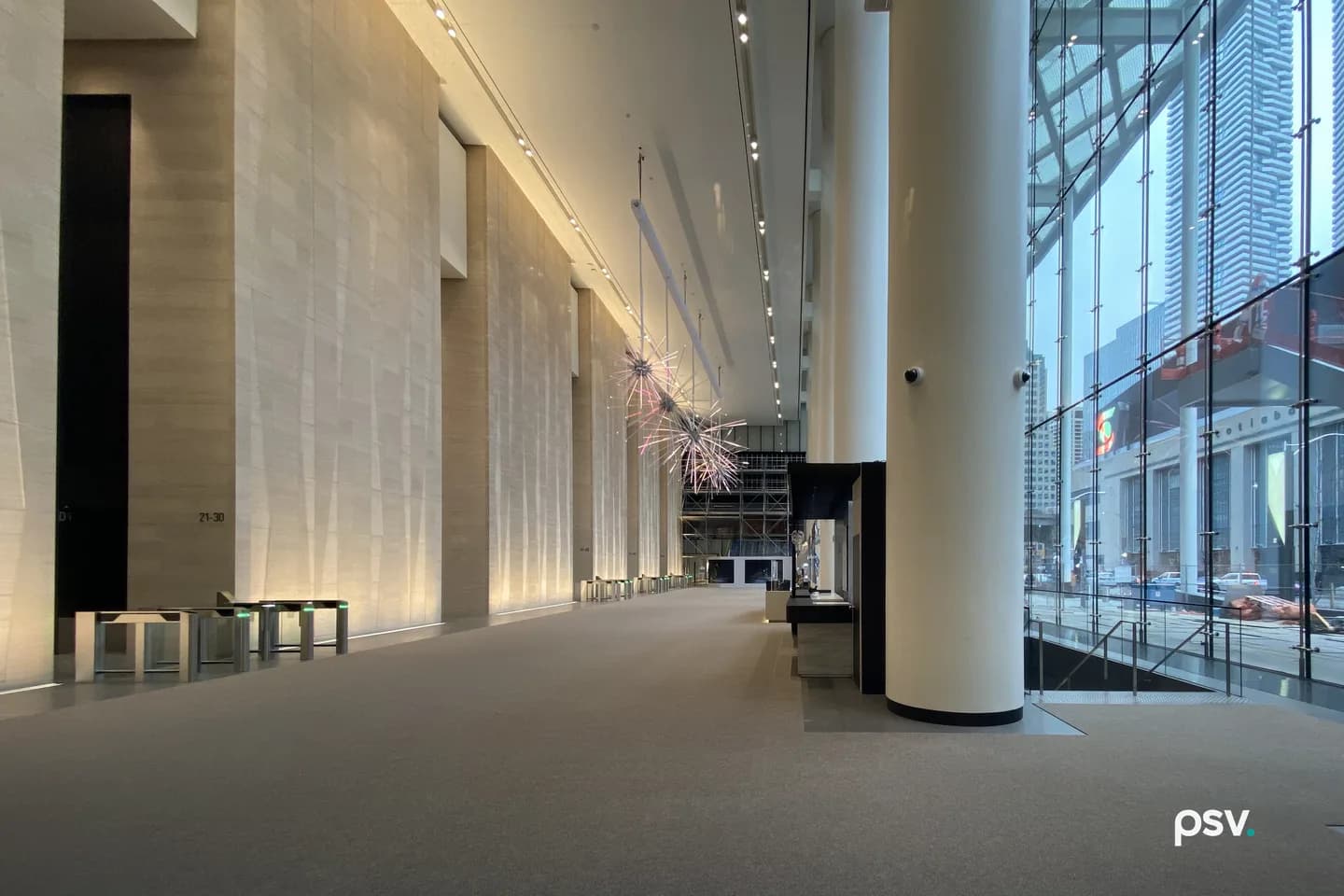 A long stone-clad office lobby with a bank of glass security speed gates glowing green along the elevator wall, a cluster of spiky starburst sculptures suspended overhead, a tall round white column at right, and a cable-braced glass curtain wall opening onto the street. A long stone-clad office lobby with a bank of glass security speed gates glowing green along the elevator wall, a cluster of spiky starburst sculptures suspended overhead, a tall round white column at right, and a cable-braced glass curtain wall opening onto the street.