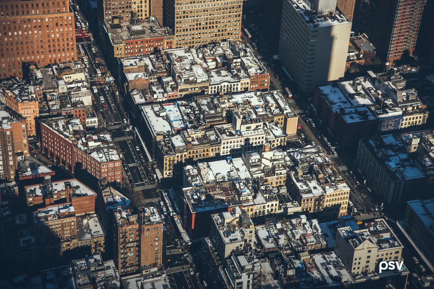 High aerial view looking down on Manhattan city blocks in low sun, showing rooftops with water tanks and mechanical units, brick and stone facades, narrow streets, and long shadows across the block interiors. High aerial view looking down on Manhattan city blocks in low sun, showing rooftops with water tanks and mechanical units, brick and stone facades, narrow streets, and long shadows across the block interiors.