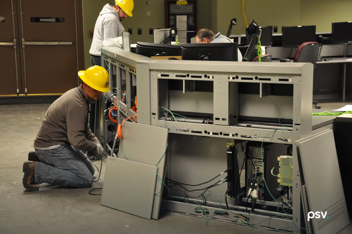 A worker in a yellow hard hat kneels to cable the open back of a grey operator console, its side panels removed and propped on the floor; a second hard-hatted worker and a seated man at monitors work behind it in a control room. A worker in a yellow hard hat kneels to cable the open back of a grey operator console, its side panels removed and propped on the floor; a second hard-hatted worker and a seated man at monitors work behind it in a control room.
