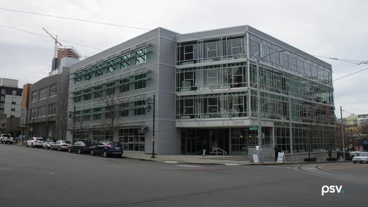 A four-story modern office building on a city street corner in Seattle, clad in silver metal panels and green-tinted glass, with ground-floor storefront windows, parked cars along the curb and overhead trolley wires.