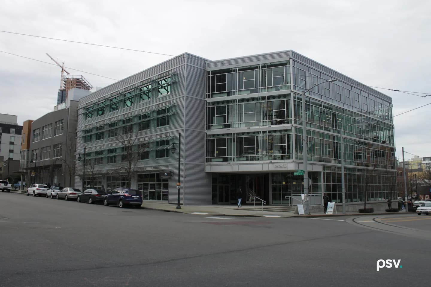 A four-story modern office building on a city street corner in Seattle, clad in silver metal panels and green-tinted glass, with ground-floor storefront windows, parked cars along the curb and overhead trolley wires. A four-story modern office building on a city street corner in Seattle, clad in silver metal panels and green-tinted glass, with ground-floor storefront windows, parked cars along the curb and overhead trolley wires.