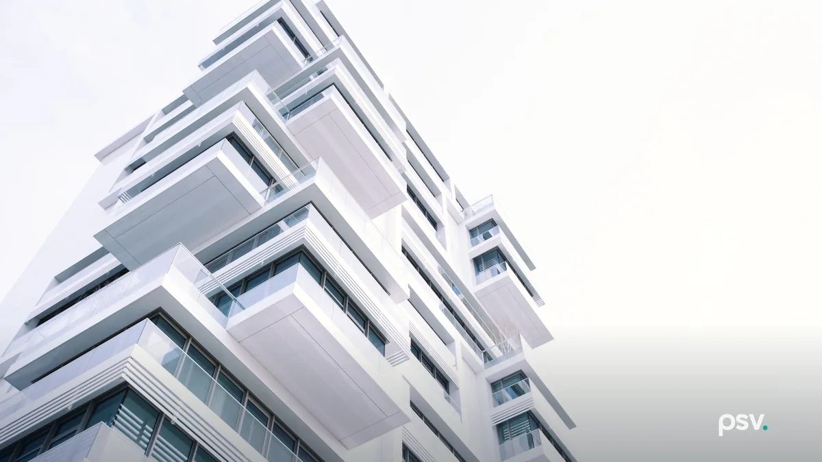 A low angle view of a white modern apartment building, its stacked projecting balconies and glass railings running diagonally against a bright overcast sky, with dark window frames and horizontal louvres on the lower floors.