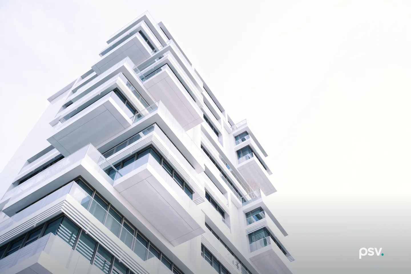 A low angle view of a white modern apartment building, its stacked projecting balconies and glass railings running diagonally against a bright overcast sky, with dark window frames and horizontal louvres on the lower floors. A low angle view of a white modern apartment building, its stacked projecting balconies and glass railings running diagonally against a bright overcast sky, with dark window frames and horizontal louvres on the lower floors.