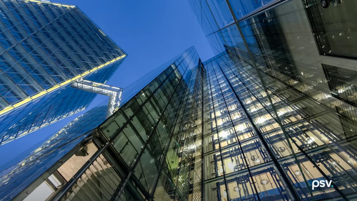 Upward view of two glass office towers at blue hour, the near facade folding at a sharp corner, with lit interior ceilings visible through the curtain wall, exposed structural framing and an enclosed walkway bridging the buildings.