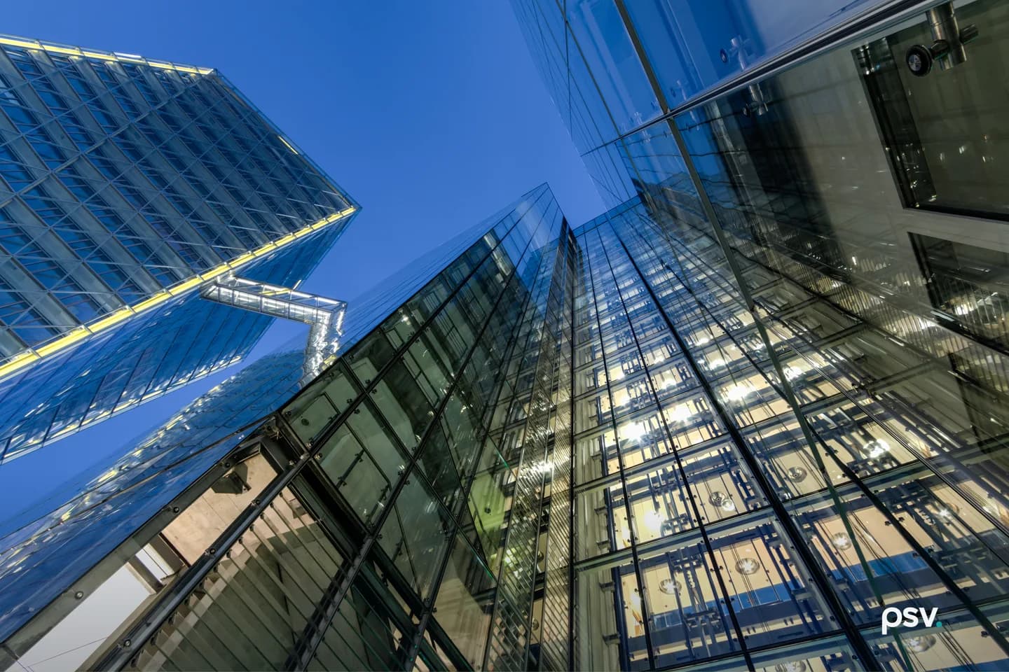Upward view of two glass office towers at blue hour, the near facade folding at a sharp corner, with lit interior ceilings visible through the curtain wall, exposed structural framing and an enclosed walkway bridging the buildings. Upward view of two glass office towers at blue hour, the near facade folding at a sharp corner, with lit interior ceilings visible through the curtain wall, exposed structural framing and an enclosed walkway bridging the buildings.