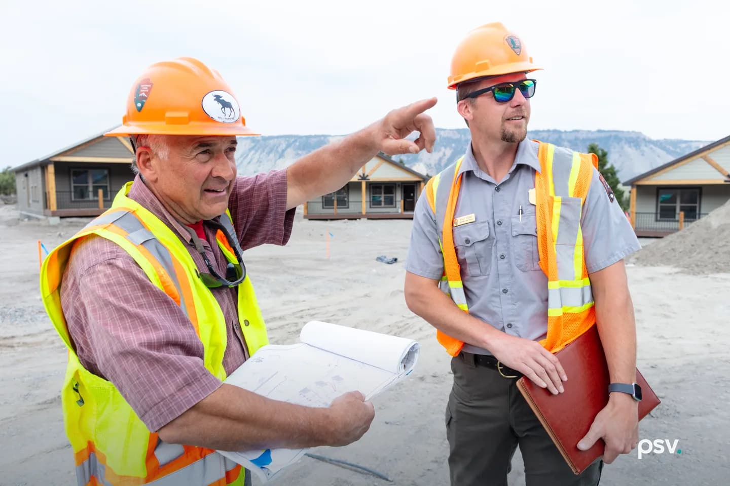 Two men in orange hard hats and reflective vests stand on a graded construction site in front of new single-story buildings. One holds an open roll of drawings and points ahead; the other carries a leather portfolio binder under his arm, listening. Two men in orange hard hats and reflective vests stand on a graded construction site in front of new single-story buildings. One holds an open roll of drawings and points ahead; the other carries a leather portfolio binder under his arm, listening.