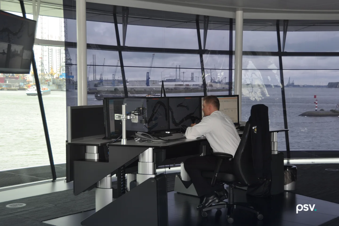 A single operator in a white uniform shirt sits at a wraparound console of flat screens showing dark river channel charts and a data panel, inside a glass-walled port control tower, with harbour cranes, two inland barges under way and a red and white channel marker beyond the windows. A single operator in a white uniform shirt sits at a wraparound console of flat screens showing dark river channel charts and a data panel, inside a glass-walled port control tower, with harbour cranes, two inland barges under way and a red and white channel marker beyond the windows.