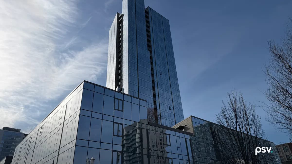 A dark blue glass residential high-rise rising behind a lower glass podium building, photographed from street level against a partly clouded sky, with bare trees and an ornate street lamp at the edges of the frame.