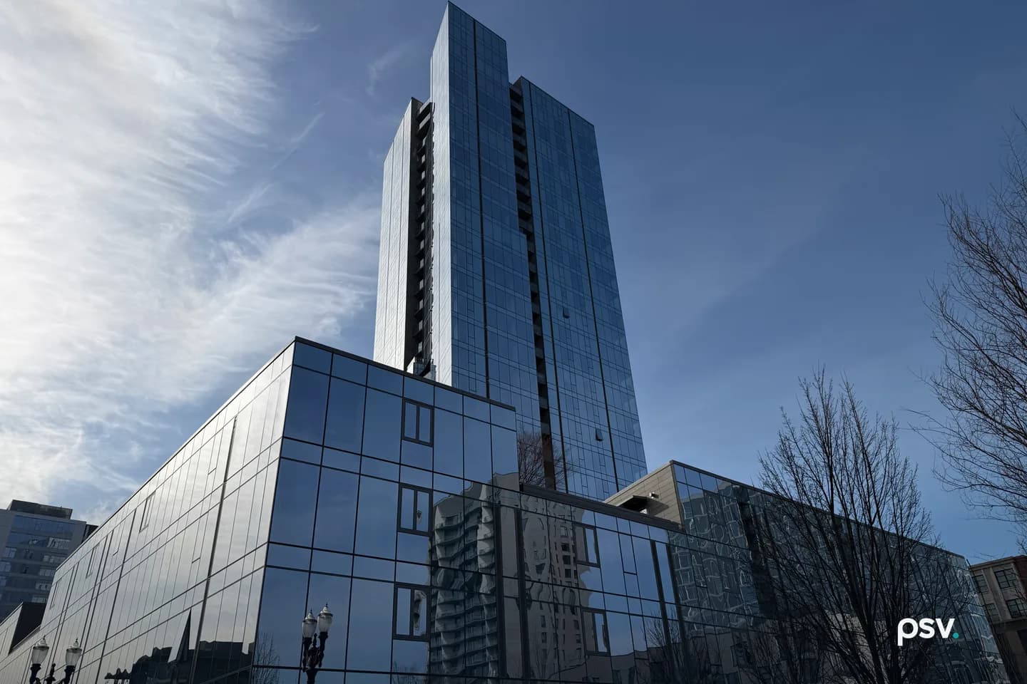 A dark blue glass residential high-rise rising behind a lower glass podium building, photographed from street level against a partly clouded sky, with bare trees and an ornate street lamp at the edges of the frame. A dark blue glass residential high-rise rising behind a lower glass podium building, photographed from street level against a partly clouded sky, with bare trees and an ornate street lamp at the edges of the frame.