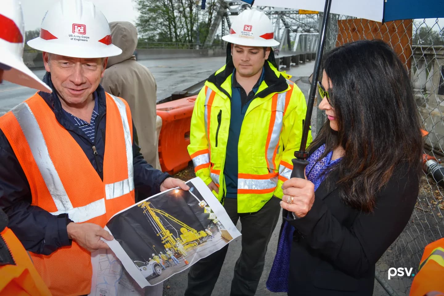 A man in a hard hat and orange safety vest holds up a printed project sheet showing a drill rig and a site map. A woman in a dark jacket studies it; a second engineer watches. A man in a hard hat and orange safety vest holds up a printed project sheet showing a drill rig and a site map. A woman in a dark jacket studies it; a second engineer watches.