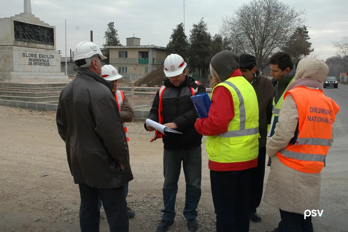 A group of engineers and agency officials in hard hats and hi-vis vests stands in a tight circle at a road construction site; the man at center holds a printed document open in both hands while the others lean in to read. A group of engineers and agency officials in hard hats and hi-vis vests stands in a tight circle at a road construction site; the man at center holds a printed document open in both hands while the others lean in to read.
