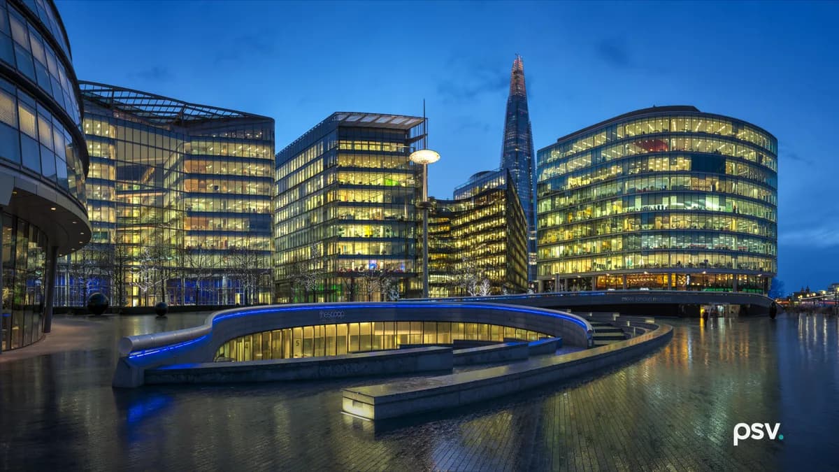 A wide dusk view of a riverside office development: a curved glass building at left, three glass and steel office blocks with lit interiors, a spired tower rising behind them, and a curved stone amphitheatre on wet reflective paving in the foreground.