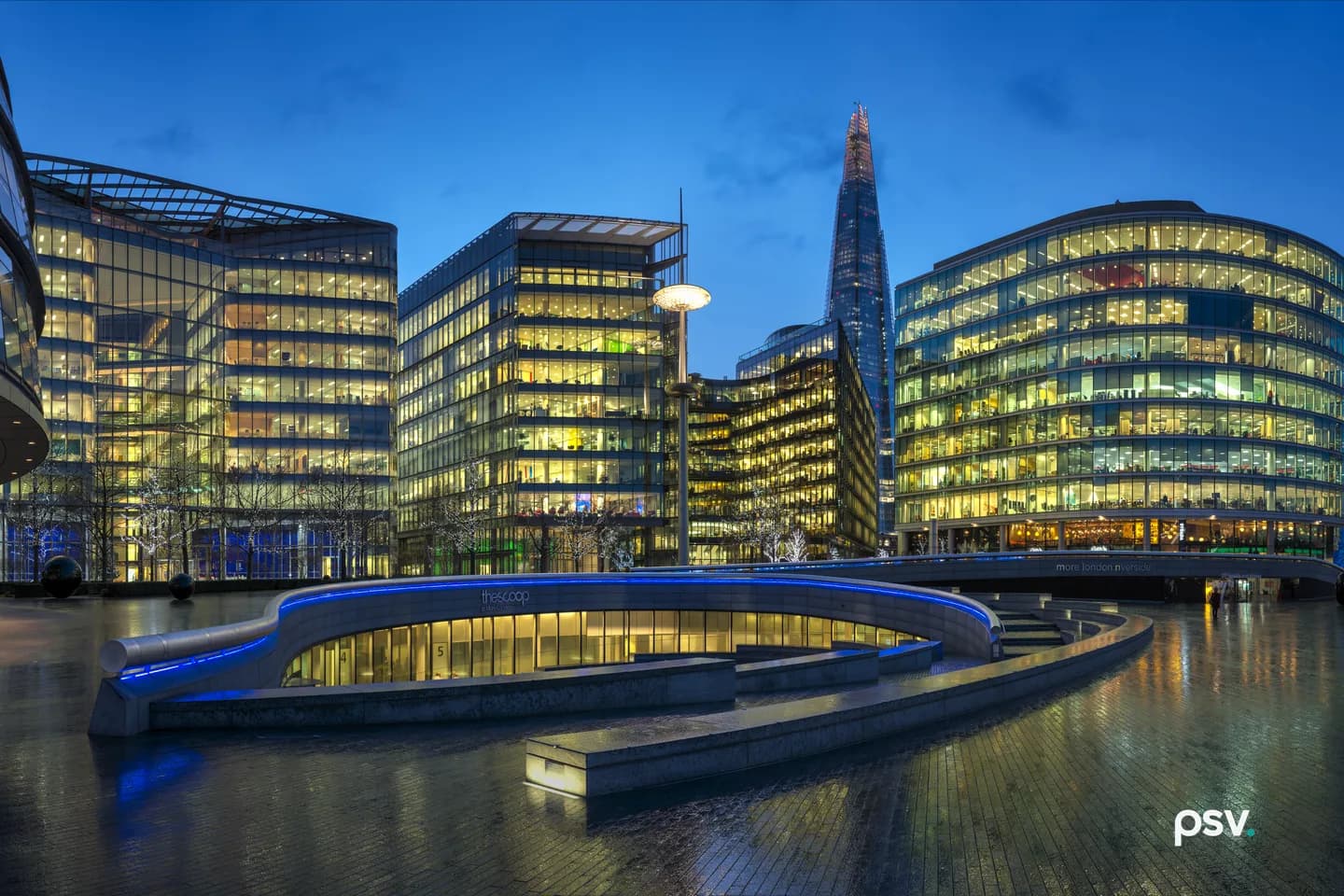 A wide dusk view of a riverside office development: a curved glass building at left, three glass and steel office blocks with lit interiors, a spired tower rising behind them, and a curved stone amphitheatre on wet reflective paving in the foreground. A wide dusk view of a riverside office development: a curved glass building at left, three glass and steel office blocks with lit interiors, a spired tower rising behind them, and a curved stone amphitheatre on wet reflective paving in the foreground.