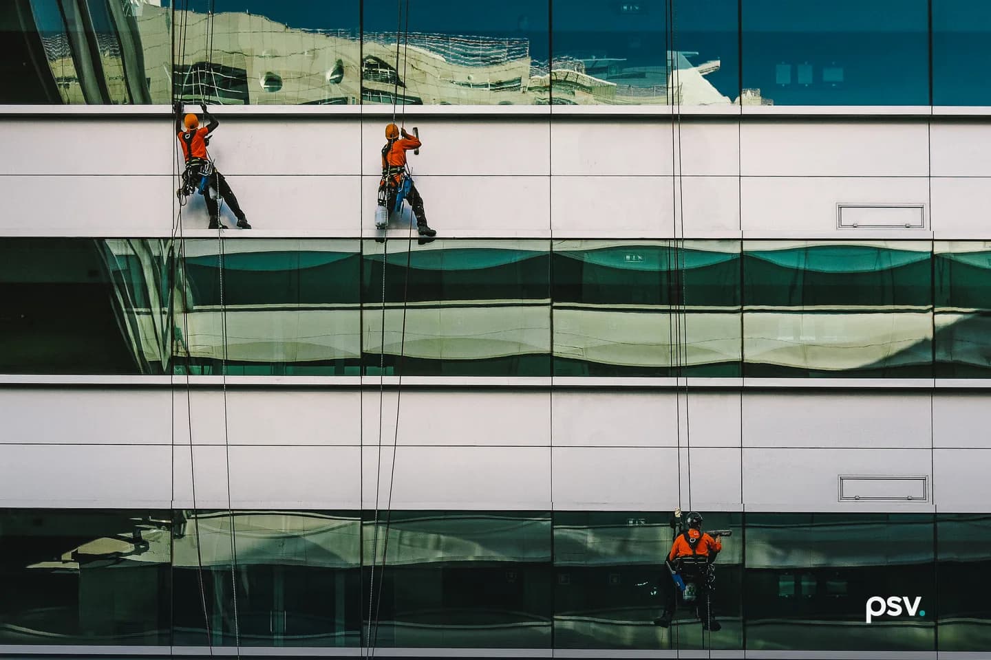 Three workers in orange shirts and helmets hang on ropes in harnesses against the glass and white panel facade of an office building, cleaning windows, with a curved building reflected in the dark green glazing. Three workers in orange shirts and helmets hang on ropes in harnesses against the glass and white panel facade of an office building, cleaning windows, with a curved building reflected in the dark green glazing.