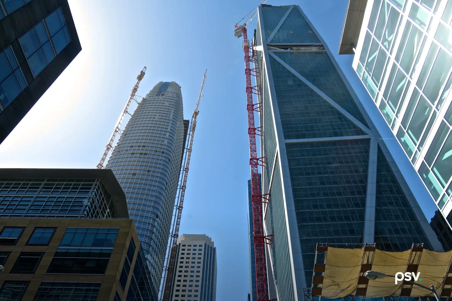 Looking straight up between San Francisco Financial District office towers on a clear day: a ribbed cylindrical high-rise still under construction with two slender tower cranes rising beside it, a faceted blue glass tower with exterior diagonal bracing on the right, a red tower crane mast running up the middle of the frame, older stone and glass office buildings across the lower left, and yellow safety netting over a sidewalk shed at the lower right Looking straight up between San Francisco Financial District office towers on a clear day: a ribbed cylindrical high-rise still under construction with two slender tower cranes rising beside it, a faceted blue glass tower with exterior diagonal bracing on the right, a red tower crane mast running up the middle of the frame, older stone and glass office buildings across the lower left, and yellow safety netting over a sidewalk shed at the lower right