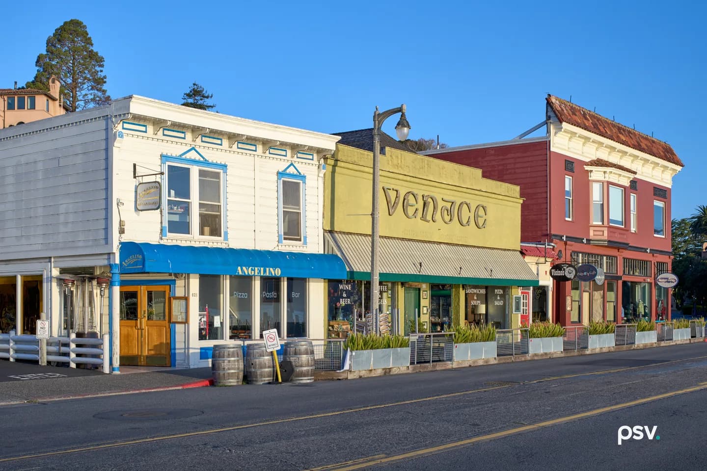 A row of low-rise storefronts on a wide empty street in early morning light: a white clapboard building with a blue awning, a yellow shopfront lettered Venice, and a red Victorian hung with tenant signs. A row of low-rise storefronts on a wide empty street in early morning light: a white clapboard building with a blue awning, a yellow shopfront lettered Venice, and a red Victorian hung with tenant signs.
