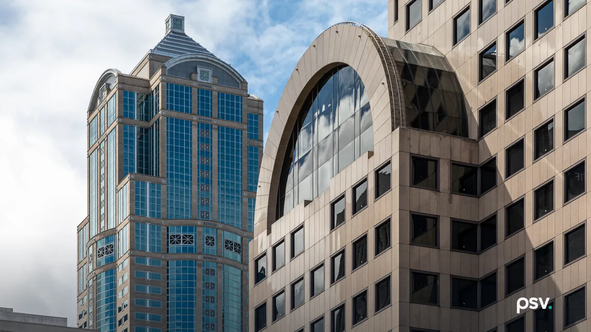 Two downtown Seattle high-rise office buildings seen from street level against a partly cloudy sky, one clad in blue-green glass with a stepped crown, the other in beige stone with a large arched window bay.