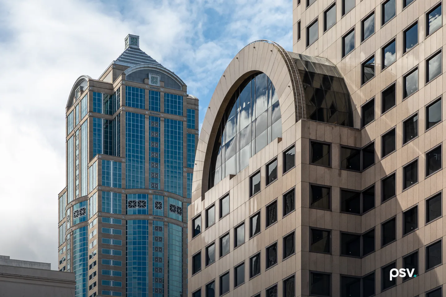 Two downtown Seattle high-rise office buildings seen from street level against a partly cloudy sky, one clad in blue-green glass with a stepped crown, the other in beige stone with a large arched window bay. Two downtown Seattle high-rise office buildings seen from street level against a partly cloudy sky, one clad in blue-green glass with a stepped crown, the other in beige stone with a large arched window bay.