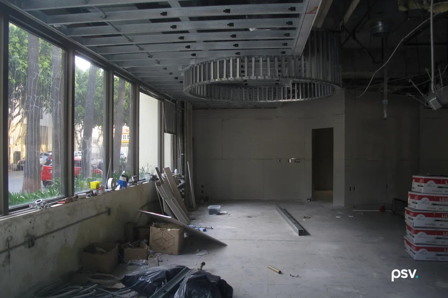 An empty commercial suite mid fit-out: a glazed storefront wall above a low concrete sill looks out to street trees and parked cars, and a metal-stud ceiling grid with a circular soffit frame hangs over bare concrete, new gypsum partitions and stacked material boxes. An empty commercial suite mid fit-out: a glazed storefront wall above a low concrete sill looks out to street trees and parked cars, and a metal-stud ceiling grid with a circular soffit frame hangs over bare concrete, new gypsum partitions and stacked material boxes.