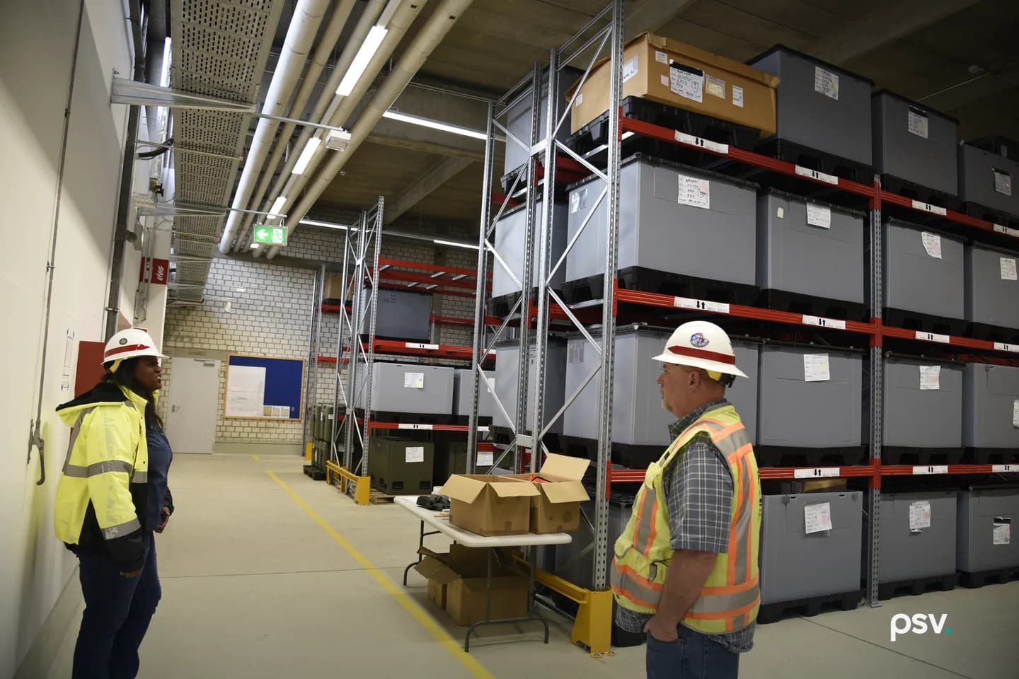 Two inspectors in high-visibility jackets and white hard hats stand facing each other in a warehouse aisle. Tall steel pallet racking loaded with labeled gray containers fills the right side, with a folding table of open cardboard boxes, yellow floor striping and exposed conduit overhead. Two inspectors in high-visibility jackets and white hard hats stand facing each other in a warehouse aisle. Tall steel pallet racking loaded with labeled gray containers fills the right side, with a folding table of open cardboard boxes, yellow floor striping and exposed conduit overhead.