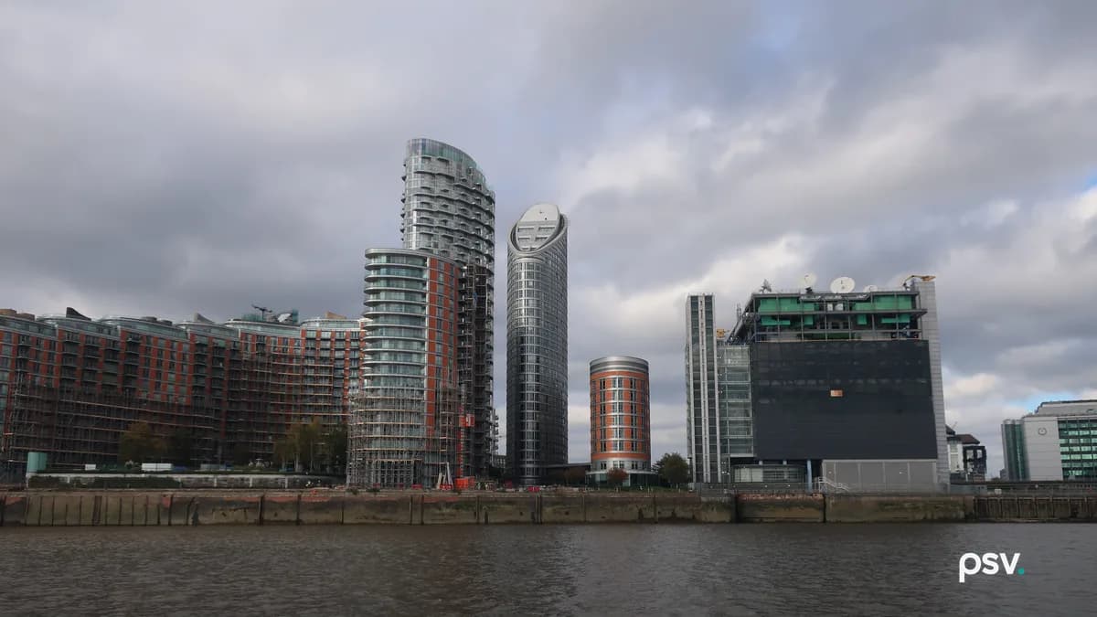 A daylight view across a river of a waterfront block in which tall residential apartment towers with balconies stand immediately beside a dark, largely windowless data centre building topped with satellite dishes and rooftop mechanical plant, with a concrete embankment along the near bank under a heavy grey sky.