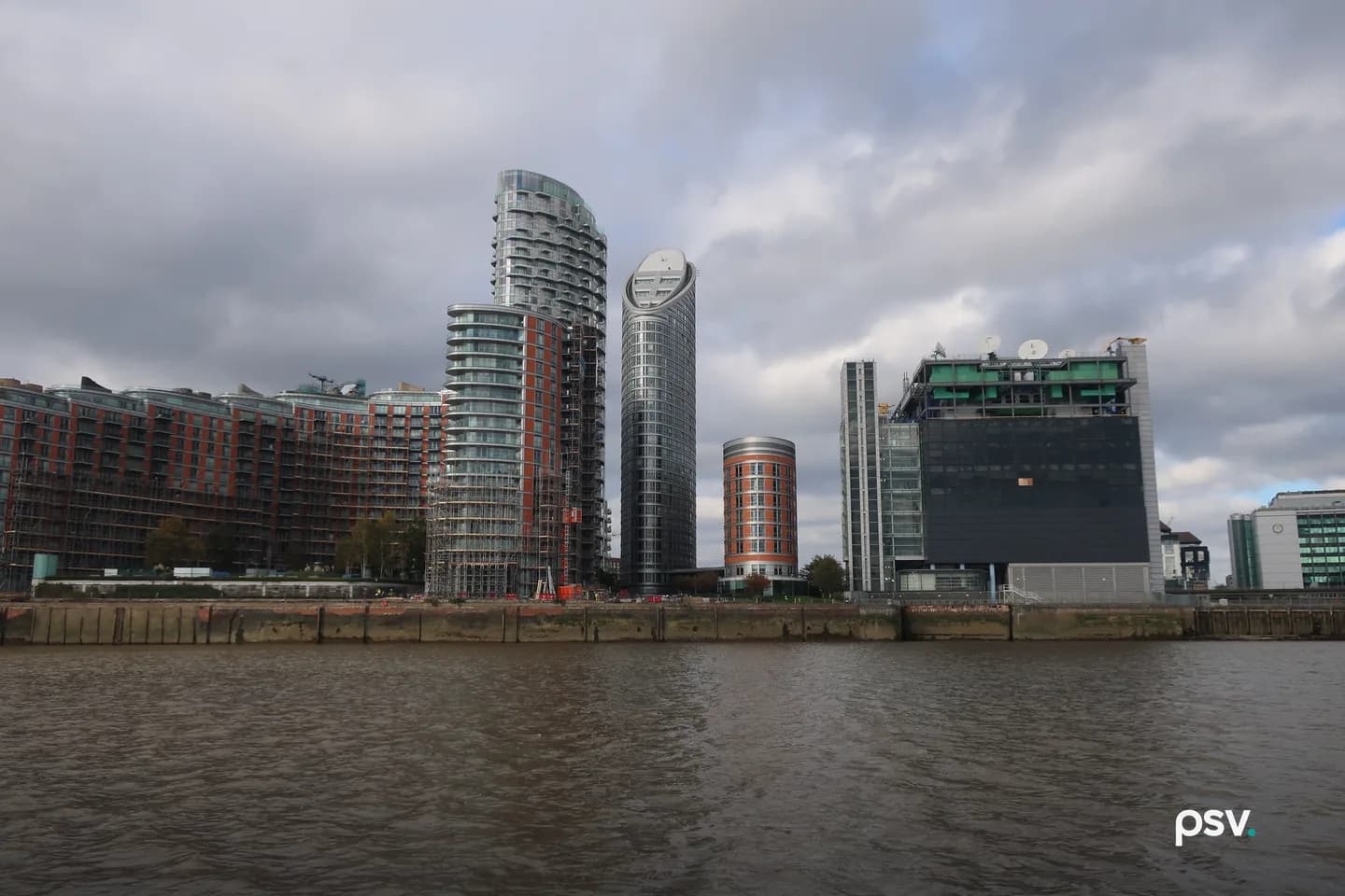 A daylight view across a river of a waterfront block in which tall residential apartment towers with balconies stand immediately beside a dark, largely windowless data centre building topped with satellite dishes and rooftop mechanical plant, with a concrete embankment along the near bank under a heavy grey sky. A daylight view across a river of a waterfront block in which tall residential apartment towers with balconies stand immediately beside a dark, largely windowless data centre building topped with satellite dishes and rooftop mechanical plant, with a concrete embankment along the near bank under a heavy grey sky.