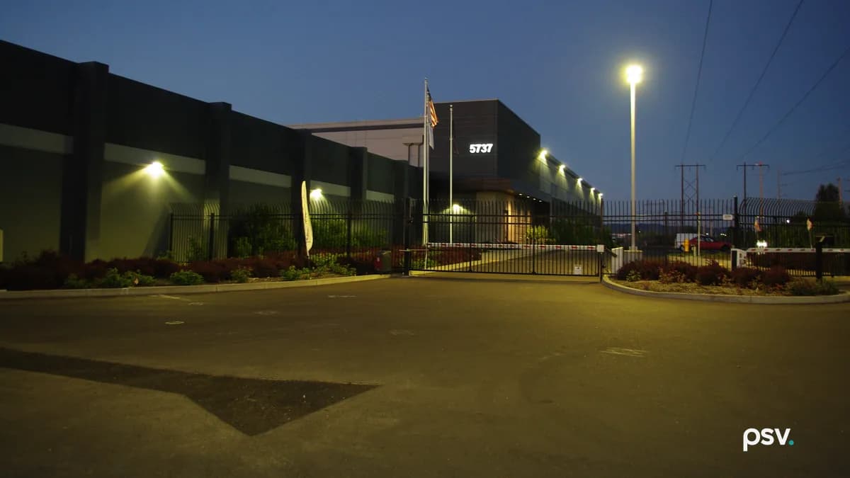 A dusk photograph of a large windowless data center building in dark grey panels behind a tall black steel security fence and a closed sliding gate with red and white barrier arms, the address number 5737 lit on the upper facade, an American flag on a pole, wall lights glowing along the building, a tall streetlight at right, high-voltage transmission towers and power lines against a deep blue sky, and an empty asphalt entry drive in the foreground.