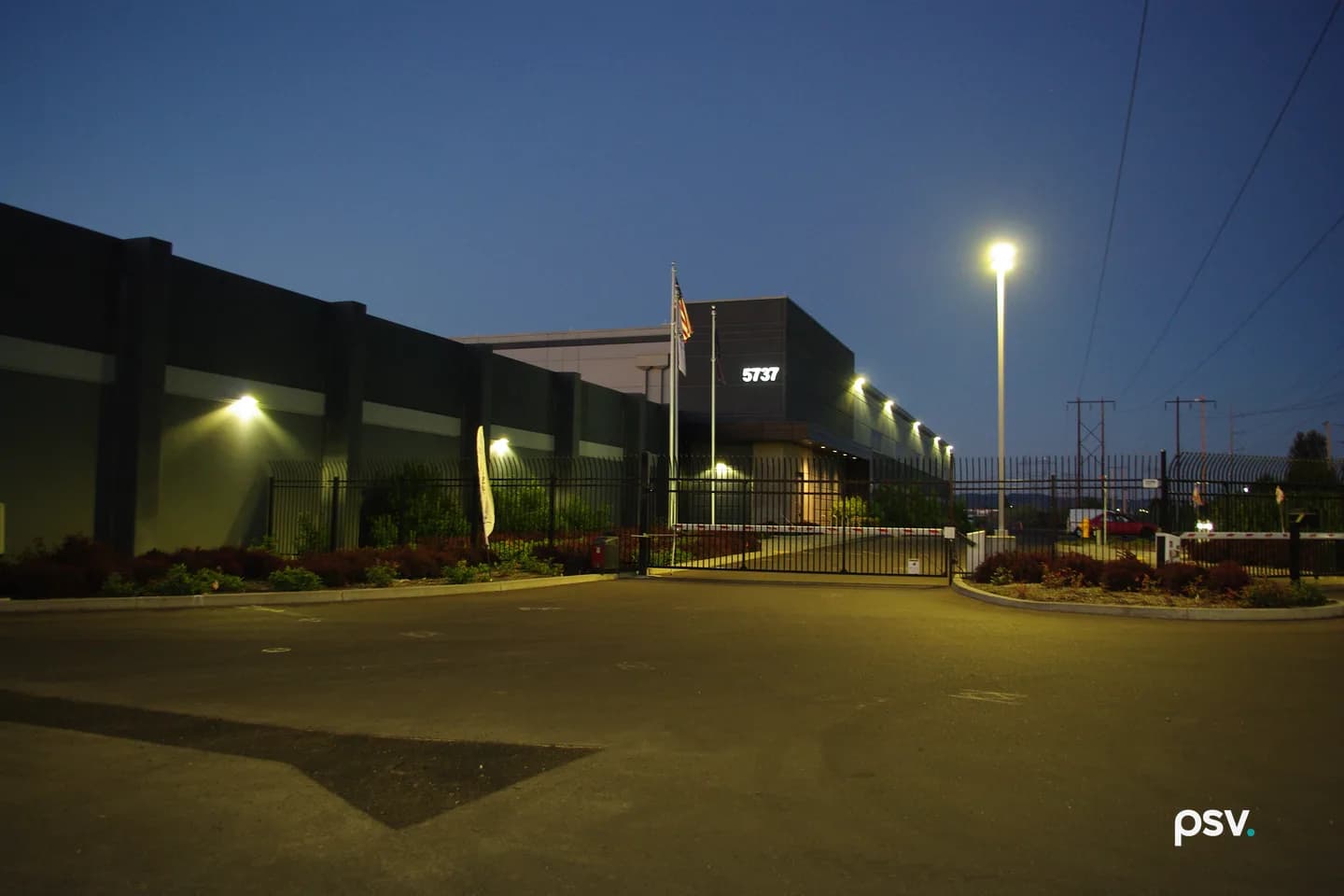 A dusk photograph of a large windowless data center building in dark grey panels behind a tall black steel security fence and a closed sliding gate with red and white barrier arms, the address number 5737 lit on the upper facade, an American flag on a pole, wall lights glowing along the building, a tall streetlight at right, high-voltage transmission towers and power lines against a deep blue sky, and an empty asphalt entry drive in the foreground. A dusk photograph of a large windowless data center building in dark grey panels behind a tall black steel security fence and a closed sliding gate with red and white barrier arms, the address number 5737 lit on the upper facade, an American flag on a pole, wall lights glowing along the building, a tall streetlight at right, high-voltage transmission towers and power lines against a deep blue sky, and an empty asphalt entry drive in the foreground.