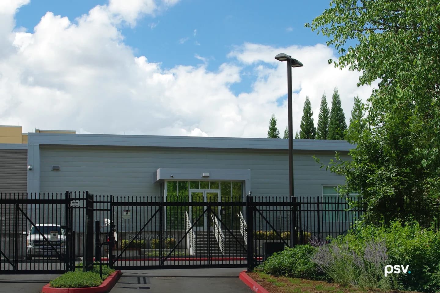A low gray-paneled data center building with a glass entrance behind a black steel security fence and closed gate, with a white pickup truck parked inside, a tall light pole, tall evergreen trees behind and landscaped shrubs in the foreground, under a blue sky with large white clouds. A low gray-paneled data center building with a glass entrance behind a black steel security fence and closed gate, with a white pickup truck parked inside, a tall light pole, tall evergreen trees behind and landscaped shrubs in the foreground, under a blue sky with large white clouds.