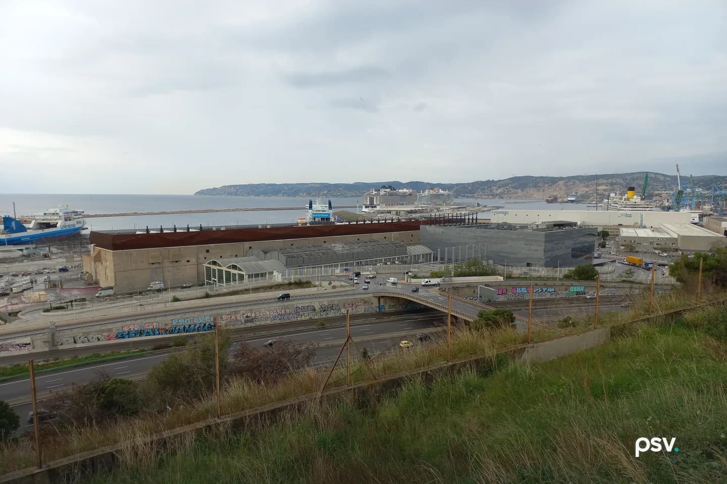 A long concrete building with a rust-colored upper storey and rows of rooftop equipment stands beside a grey metal-clad building at the edge of a harbor, with a highway and graffiti-covered walls in front, a grassy slope and wire fence in the foreground, and ferries, cruise ships, port cranes and a rocky headland under an overcast sky behind. A long concrete building with a rust-colored upper storey and rows of rooftop equipment stands beside a grey metal-clad building at the edge of a harbor, with a highway and graffiti-covered walls in front, a grassy slope and wire fence in the foreground, and ferries, cruise ships, port cranes and a rocky headland under an overcast sky behind.