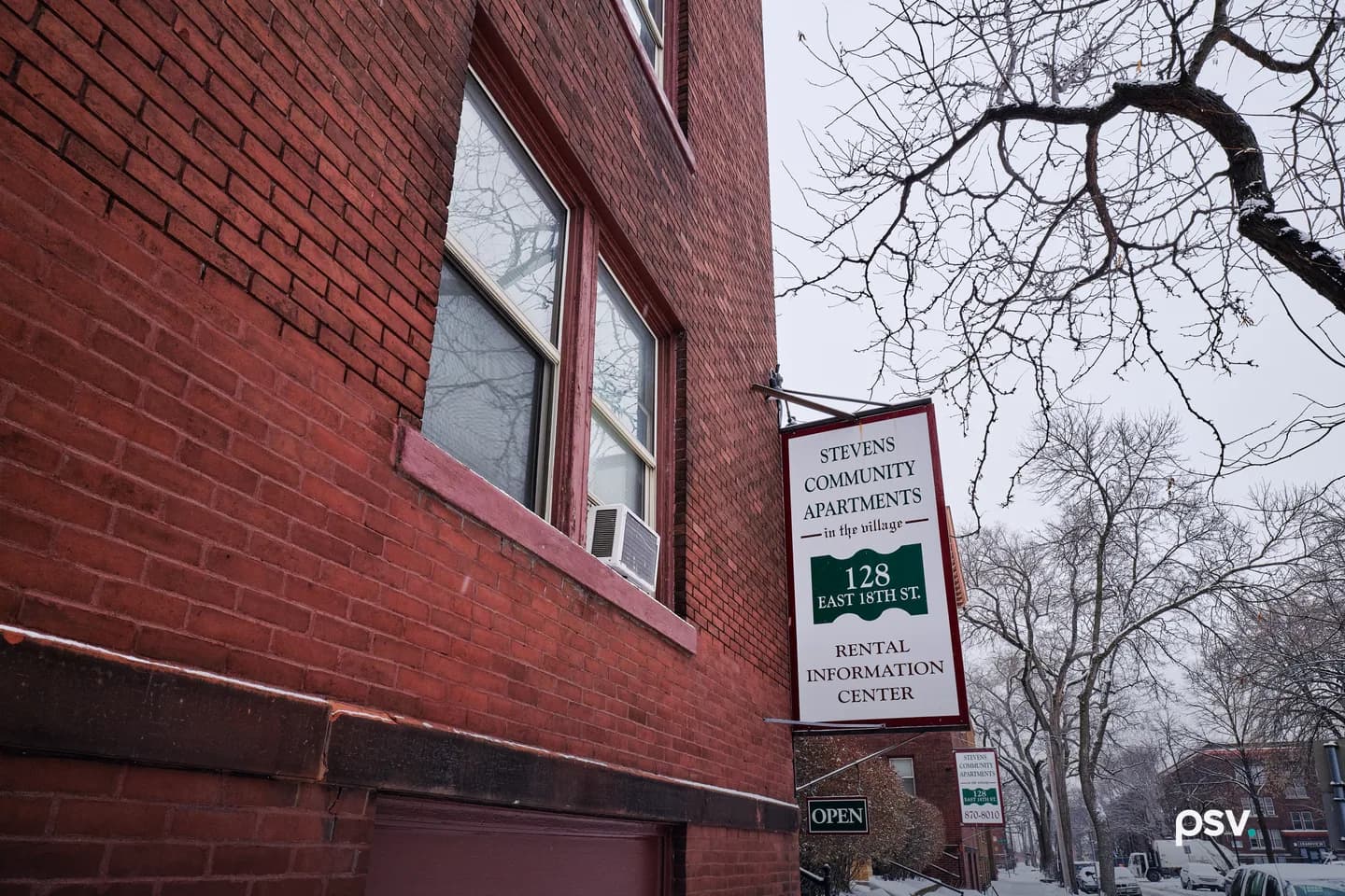 A daylight winter photograph looking up at the corner of a red brick apartment building, with double-hung windows and a window air conditioner, and a hanging sign reading Stevens Community Apartments in the village, 128 East 18th St., Rental Information Center, above a small Open sign, with bare trees, a snowy sidewalk and parked cars along the street. A daylight winter photograph looking up at the corner of a red brick apartment building, with double-hung windows and a window air conditioner, and a hanging sign reading Stevens Community Apartments in the village, 128 East 18th St., Rental Information Center, above a small Open sign, with bare trees, a snowy sidewalk and parked cars along the street.